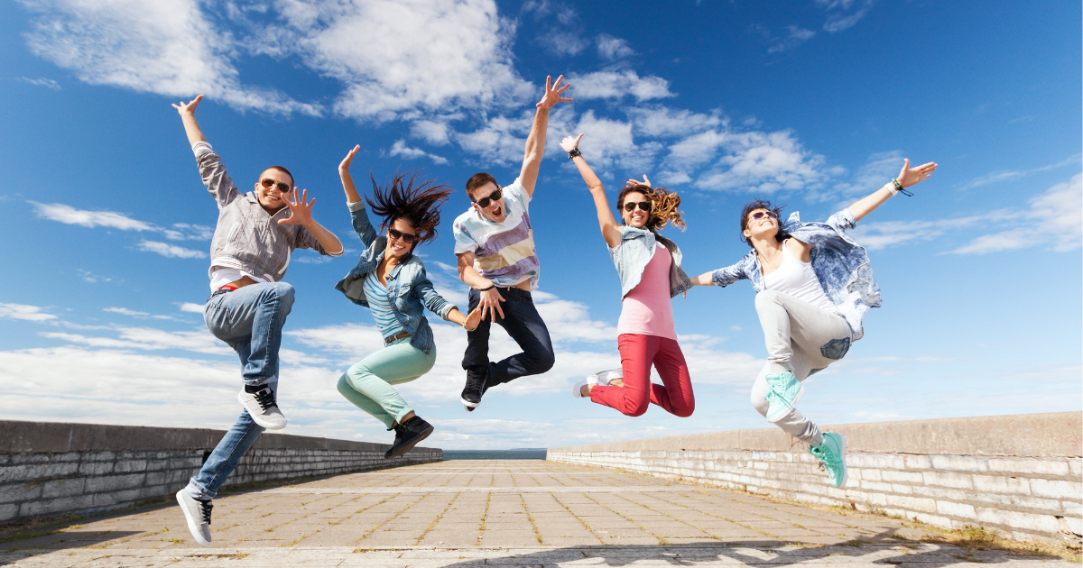 A photo of five diverse--gender and race--youths pictured mid-leap against a backdrop of blue sky and some clouds. They are on a cement path or road and they all look super excited.