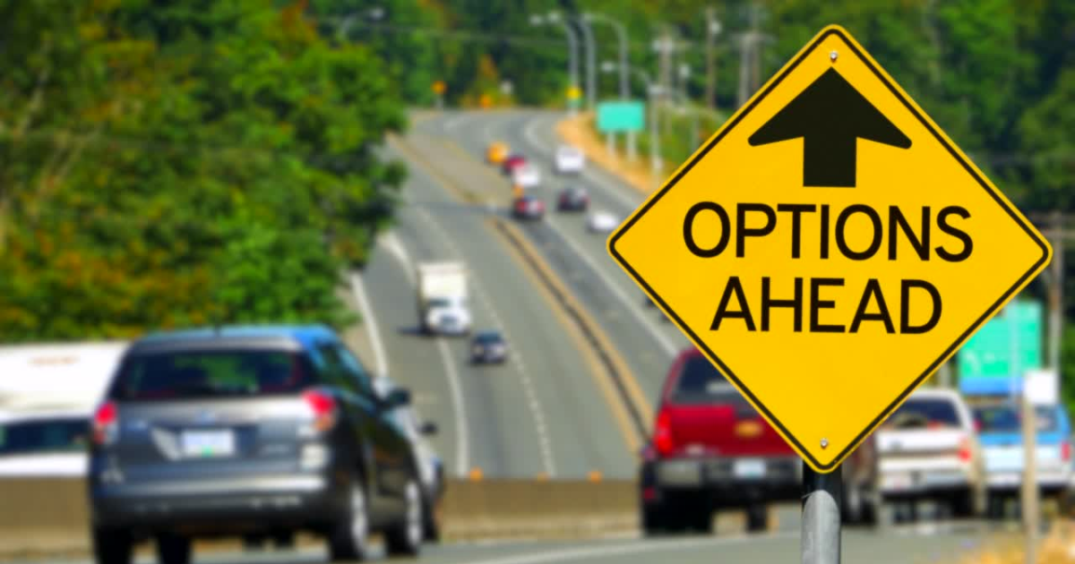 The image depicts a yellow diamond-shaped road sign in the foreground with a bold black arrow pointing upward. Beneath the arrow, in large capital letters, are the words "OPTIONS AHEAD." The sign is positioned beside a multi-lane road with several cars traveling in both directions. The background features a lush, green landscape with trees lining the road, and the road itself slightly curves into the distance.